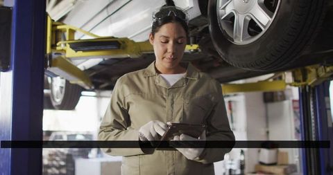 Female mechanic inspecting tablet under car on lift in auto repair workshop