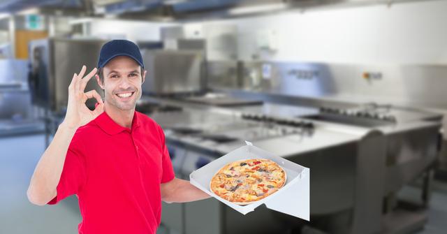 Smiling delivery man in a red uniform holds a pizza box and shows an OK gesture while standing in a commercial kitchen. Ideal for use in food delivery service marketing, restaurant advertisements, hospitality industry promotions, and culinary business websites.