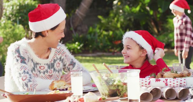 Family enjoying a Christmas meal outside in sunny weather, all wearing Santa hats and smiling. Perfect for holiday-themed advertisements, festive greeting cards, or social media posts celebrating family happiness during Christmas.