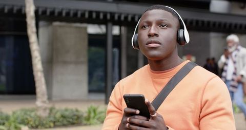 African american student listening to music with headphones holding phone on urban campus