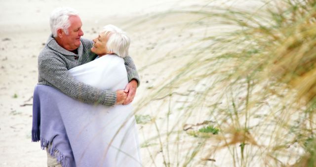 Senior couple hugging under blanket on sandy beach. Ideal for content on love, bonding, elderly lifestyle, outdoor activities, and happy retirement. Perfect for ads, articles, and blog posts about family values, senior health, and enjoying nature in older age.