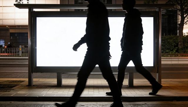 Backlit urban silhouettes walking past illuminated bus shelter billboard on city night street