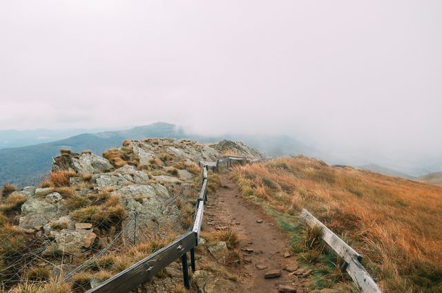 Mountain trail in foggy weather with rocky path and rustic wooden railings. Ideal for use in travel blogs, nature websites, and adventure magazines to evoke a sense of exploration and serenity in the wild. It shows elements of hiking, trekking, and the beauty of remote landscapes.