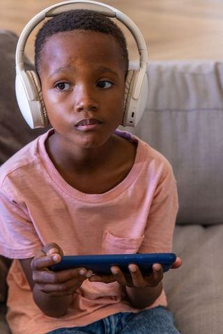 African american child wearing headphones holding smartphone on couch, listening and streaming