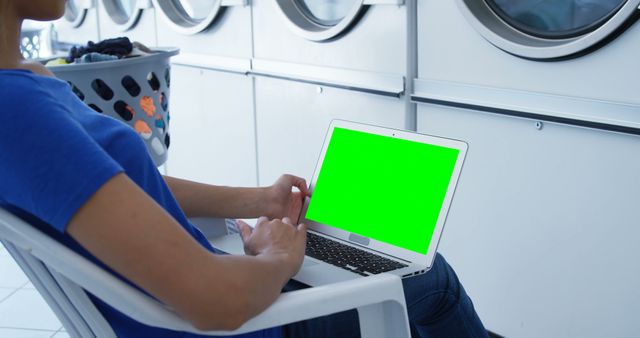 Person is sitting in a laundry room using a laptop with a green screen for easy customization. Background shows washing machines, indicating a busy yet productive setting. Useful for depicting multitasking, remote work, or technology integration in daily life. Perfect for advertisements, blogs, or articles about modern lifestyle, technology, productivity, or household chores.