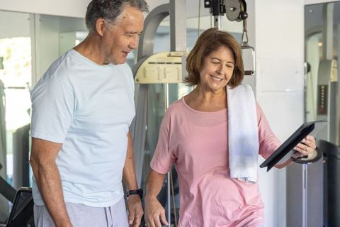 Senior couple checking tablet at gym, woman smiling with towel after workout session