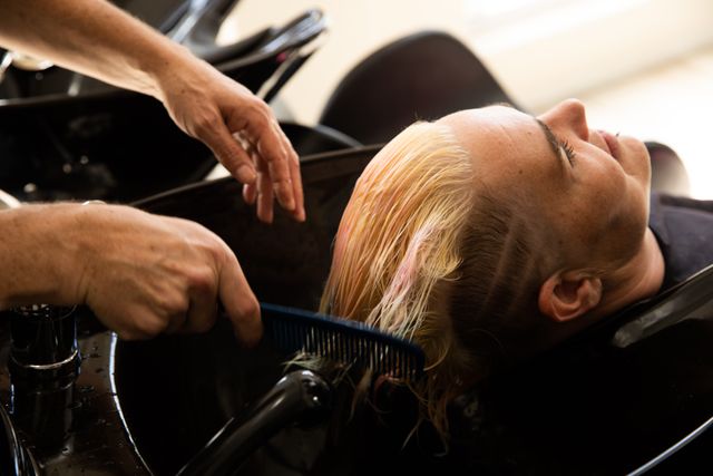 Side view of a middle aged biracial female hairdresser and a young Caucasian woman having her hair washed and brushed with a comb in a hair salon