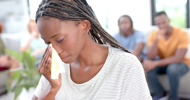 Young woman wiping her tears during a group therapy session with people offering support in the background. Perfect for use in articles or content focused on mental health, emotional well-being, group therapy, supportive communities, and personal stories.