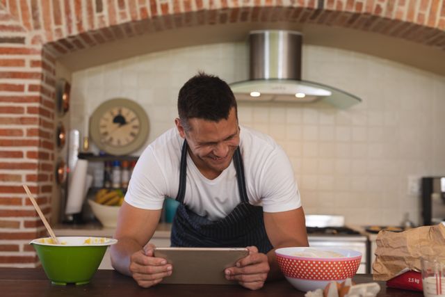 Front view of a caucasian man leaning on a kitchen table while holding a tablet with a grin on his face. beside him are cooking ingredients.