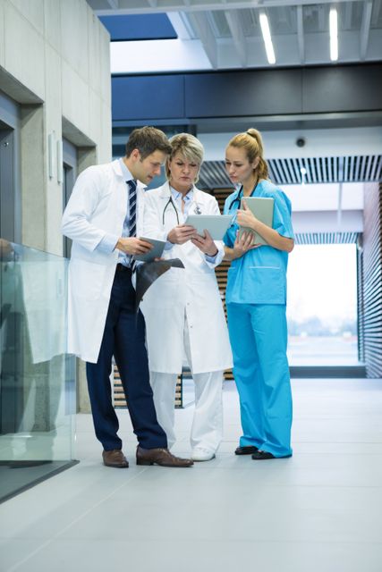 Medical team consisting of doctors and a nurse discussing patient information using a digital tablet in a hospital corridor. Ideal for use in healthcare, medical technology, teamwork, and hospital-related content.