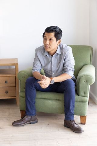 Mid-adult asian man sitting on green armchair, contemplative casual style in minimalist interior