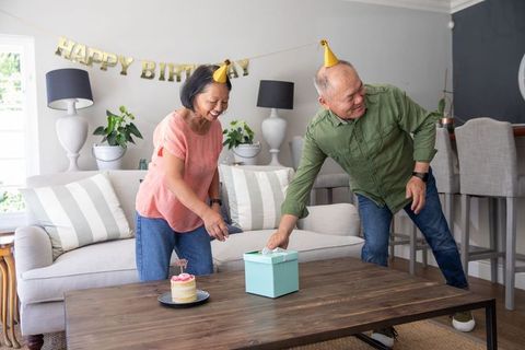 Senior asian couple celebrating birthday at home wearing party hats while opening gift