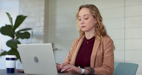 Young professional woman typing on laptop in coworking space with coffee and smartphone