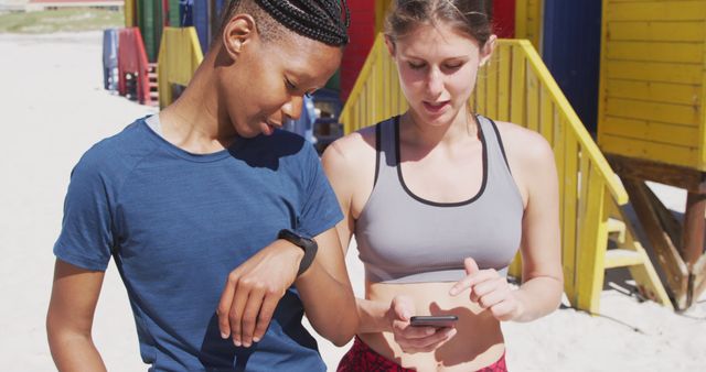 Two women in sportswear checking fitness data on a smartwatch at a beach, indicating an active and healthy lifestyle. This can be used for advertisements, lifestyle blogs, fitness promotion, or technology use in daily life.