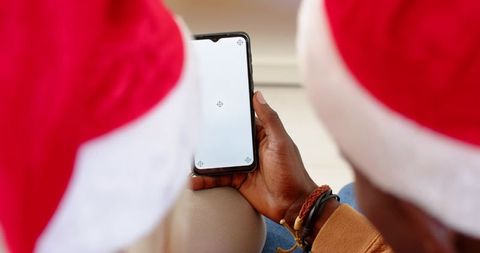 Diverse couple wearing santa hats sharing smartphone with blank screen for holiday mockup