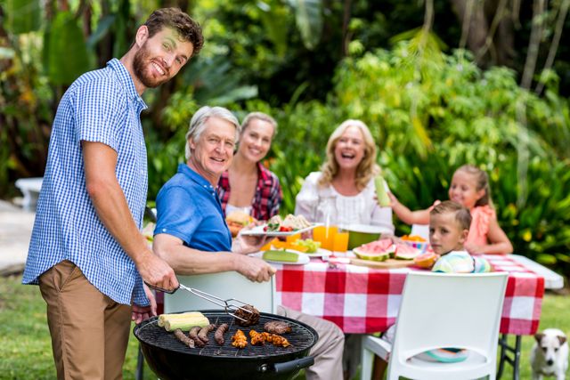 Multigenerational family enjoying a barbecue in a lush backyard. Father grilling food while family members, including grandparents and children, sit at a table with a red and white checkered tablecloth. Perfect for depicting family gatherings, outdoor activities, and summer leisure. Ideal for use in advertisements, blogs, and articles about family time, outdoor cooking, and summer events.