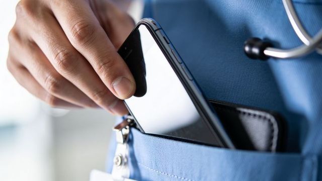 Nurse sliding smartphone into blue scrub pocket with stethoscope and id badge