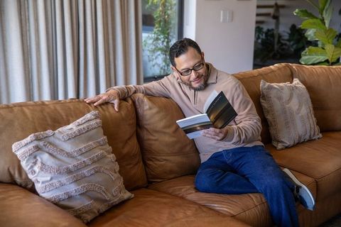 Middle-aged asian man reading paperback on leather sofa in modern cozy living room