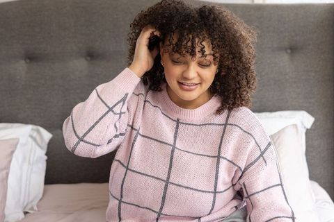 Smiling woman relaxing on bed in pink knit sweater touching curly hair, cozy bedroom