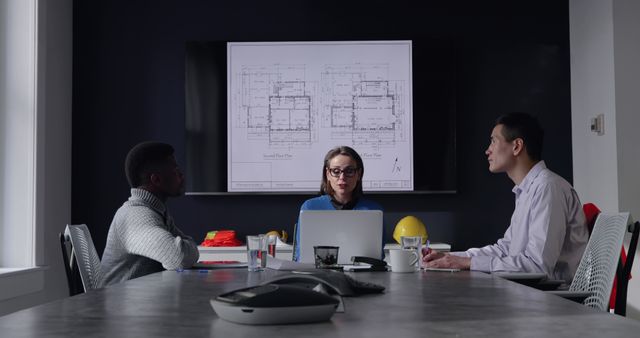 The image shows a group of architects sitting around a conference table, reviewing building plans displayed on a screen. A laptop and various documents are on the table, indicating an ongoing discussion. This image is ideal for use in articles or presentations about professional teamwork, architecture, business meetings, project planning, and collaborative efforts within the workplace.