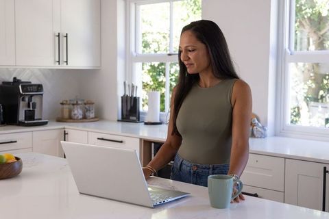 Indian woman working on laptop at modern sunlit kitchen island holding mug remote work