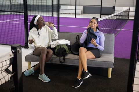 Diverse female teammates taking break on purple court sofa, drinking water