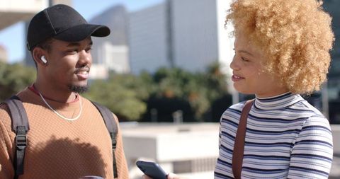 Young diverse friends chatting on sunlit urban plaza holding smartphone and backpacks