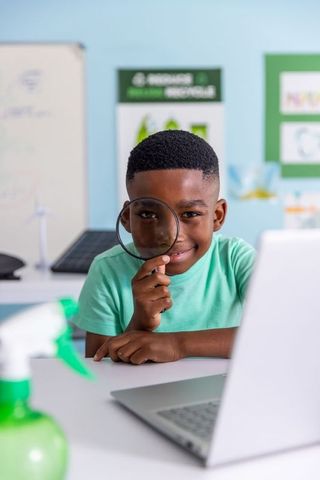 African american boy exploring clean energy with magnifying glass and laptop in classroom