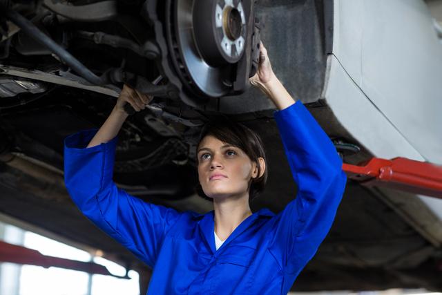 This image shows a female mechanic working on a car's wheel disc brake in a repair garage. Suitable for use in articles and content related to women in the automotive industry, car maintenance and repair services, gender diversity in the workplace, and promoting skilled trades careers. Ideal for business brochures, websites, and marketing materials for car repair shops and technical training programs.