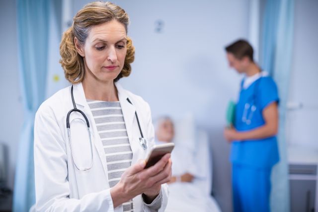 Female doctor standing in hospital room, using mobile phone while a patient rests in the background with a nurse attending them. Useful for illustrating concepts related to healthcare, medical professionals, technology use in medicine, hospital communication, and modern healthcare settings.