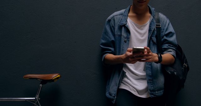 Teenager standing against a dark wall, wearing a denim jacket and a backpack, texting on a smartphone. Bicycle partially visible to the side. Suitable for topics on modern youth culture, technology usage, urban lifestyle, and casual fashion.