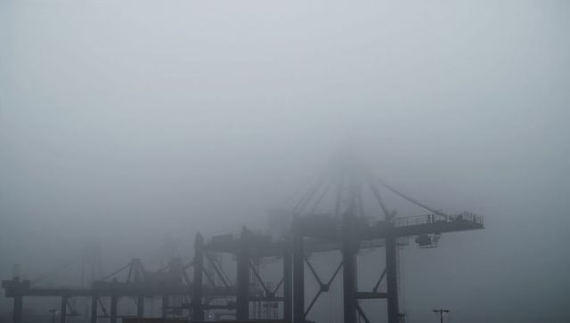 Gantry cranes emerging from dense fog at harbor terminal, moody industrial skyline silhouettes