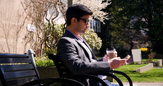 A young Asian businessman in a suit is sitting on a park bench using his smartphone, with copy space. He appears focused on his device, managing work tasks while enjoying a sunny outdoor setting.