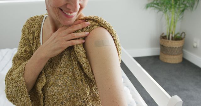 Middle-aged woman smiling and showing arm with bandage after getting vaccinated at home, suggesting health and wellness, suitable for campaigns promoting vaccination, medical blogs, or health-related articles.