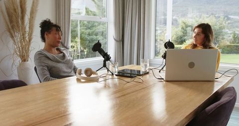 Diverse women cohosting home podcast at wooden table with laptop and microphones