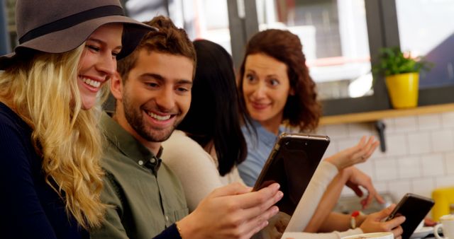 Group of young friends chatting and enjoying social media on their mobile phones while seated at a cafe. They are smiling and engaging with each other, representing connectivity through technology. Perfect for uses related to socializing, modern communication, youth culture, technology in everyday life, and cafe or restaurant advertising.