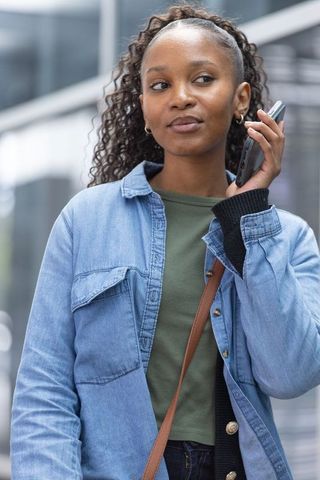 Young woman using smartphone outside glass office wearing denim jacket and crossbody bag