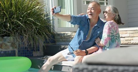Asian senior couple taking selfie at sunny backyard pool, enjoying relaxed summer moment