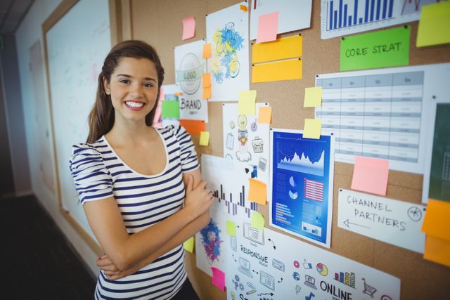 Young female executive standing confidently with arms crossed near a bulletin board filled with charts, graphs, and planning materials. Ideal for use in business, corporate, and teamwork-related content, showcasing professional environments and strategic planning.