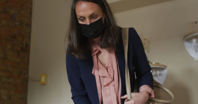Woman in indoor environment wearing a black face mask, dressed in professional attire and carrying a beige bag. Perfect for use in articles about pandemic precautions, health and safety, business continuity during health crises, and general hygiene practices.
