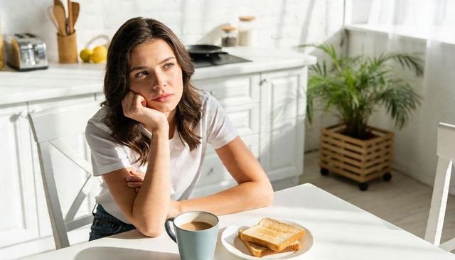 Young woman sitting at breakfast table gazing thoughtfully in sunlit minimalist kitchen