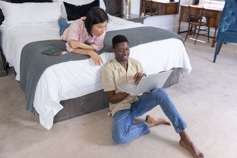 Asian and african american couple planning trip on laptop in cozy bedroom with passport
