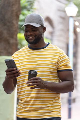 African man standing on city street smiling while checking phone and holding coffee cup