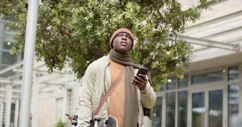 African american student holding phone, skateboard beneath urban canopy wearing beanie scarf