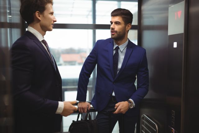 Two businessmen in formal attire are conversing in an office elevator. One is holding a briefcase, suggesting a professional setting. This image can be used for business-related content, corporate communications, workplace interactions, and professional networking themes.