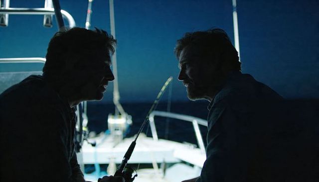 Two fishermen sharing quiet conversation on boat stern at night, fishing rod in hand