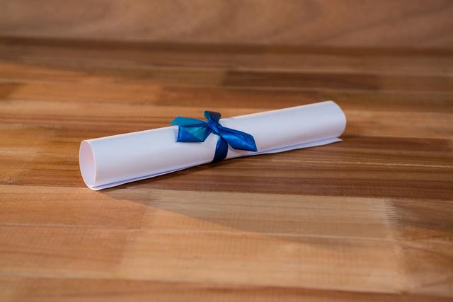 Close-up of a graduation certificate rolled and tied with a blue ribbon, placed on a wooden table. Ideal for use in educational materials, graduation announcements, academic achievement promotions, and celebratory content.