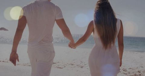 Romantic Couple Walking Hand in Hand on Sunlit Beach Wearing White Outfits Back View