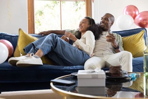 Couple relaxing on blue sofa holding wine glasses amid balloons and gift celebration