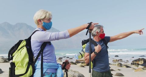 Senior couple hiking on beach with face masks and backpacks